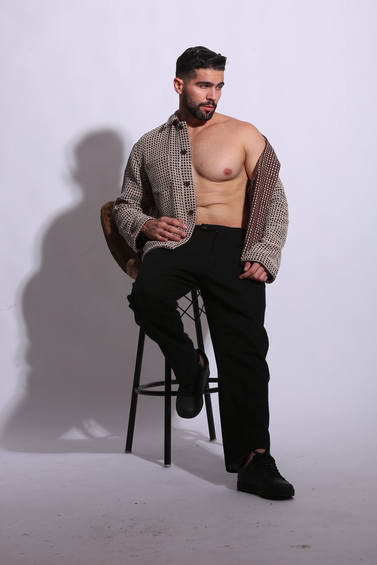 Man sitting on a chair without a shirt, wearing a patterned jacket and black pants.