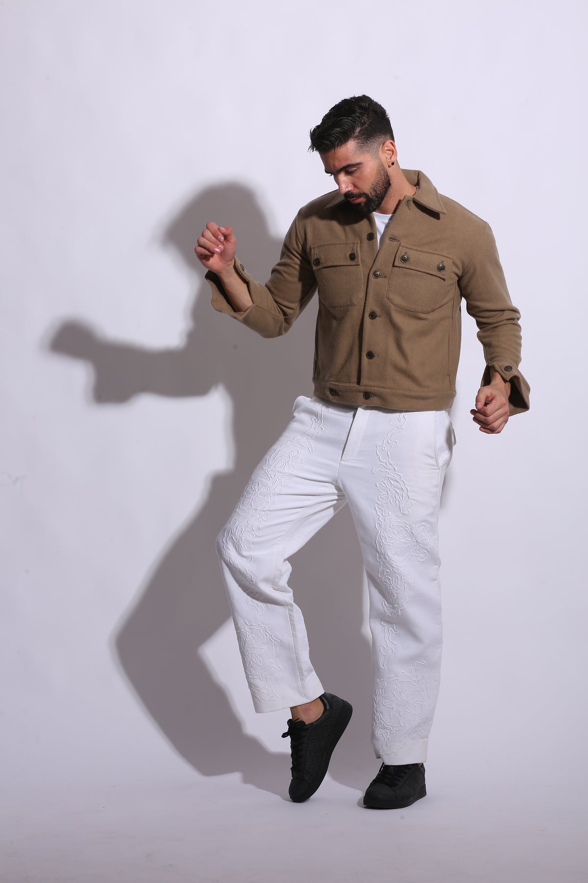 Man in a trench coat and white pants standing on a white background with a shadow effect.