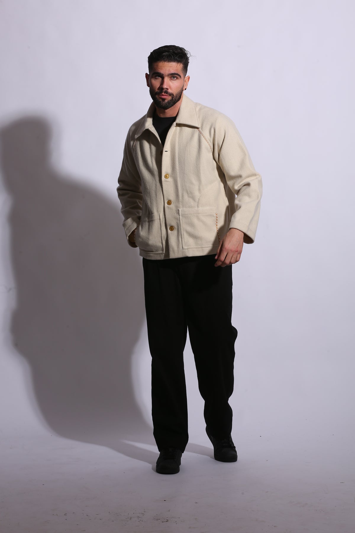 Person wearing a beige jacket and black pants standing on a white floor with a shadow cast behind them.