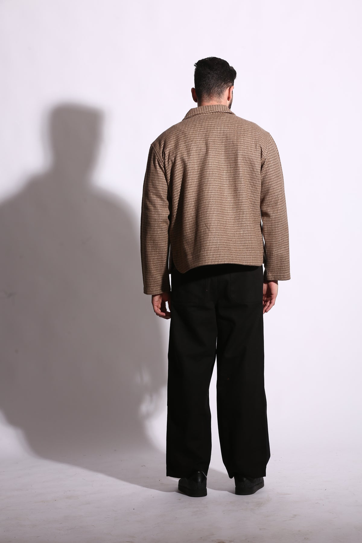 Person wearing a brown coat and black pants standing on a white floor with a shadow cast on the wall.