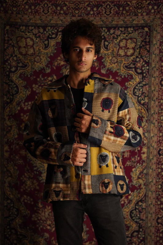 A person standing in front of a patterned backdrop wearing a multicolored patchwork shirt jacket with sheep applique.