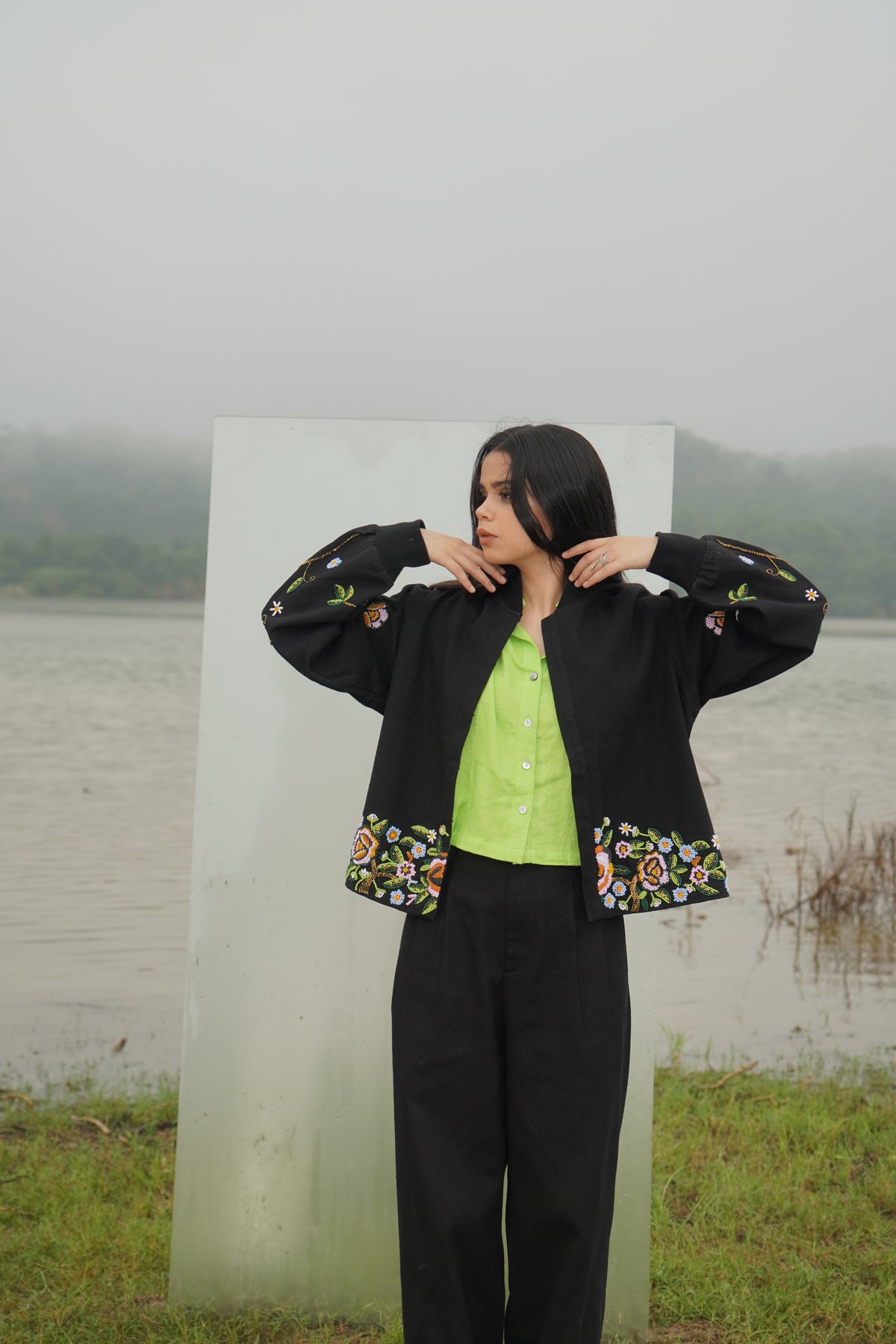 A person wearing a black unisex jacket with floral embellishments and a green top, standing near a transparent panel with a natural background.