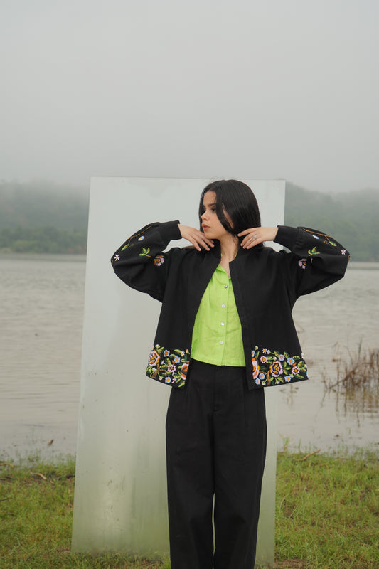 A person wearing a black unisex jacket with floral embellishments and a green top, standing near a transparent panel with a natural background.